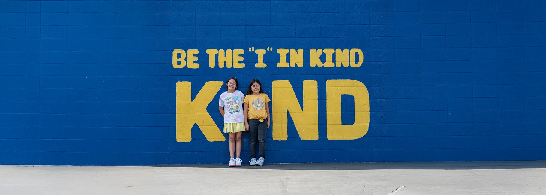 Two AESD students stand between the letters of 'KIND' on a blue wall, symbolizing the message Be the I in KIND.